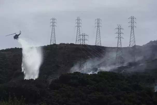 FILE - In this Oct. 10, 2019, file photo, a helicopter drops water near power lines and electrical towers while working at a fire on San Bruno Mountain near Brisbane, Calif. California energy leaders on Friday, May 6, 2022 said the state may see an energy shortfall this summer. Threats from drought, extreme heat and wildfires, are among the issues that will create challenges for energy reliability this summer and in the coming years. (Credit: AP Photo/Jeff Chiu, File)