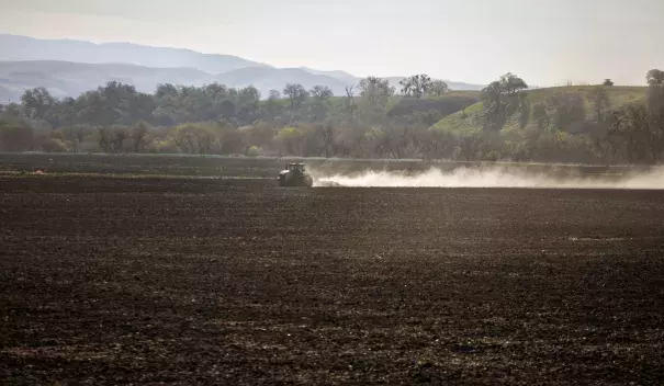 Image shows a field with a tractor plowing and stirring up dust.