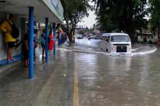 A woman stands on a bus stop bench as a driver of a Volkswagen van navigates a flooded street in Recife, state of Pernambuco, Brazil, Saturday, May 28, 2022. (Credit: AP Photo/Marlon Costa/Futura Press)
